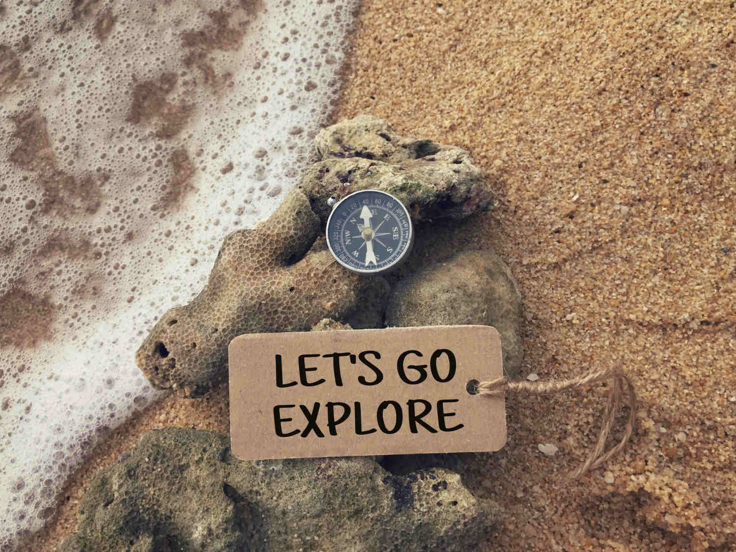 Schriftzug 'Let's Go Explore' mit Kompass am Strand